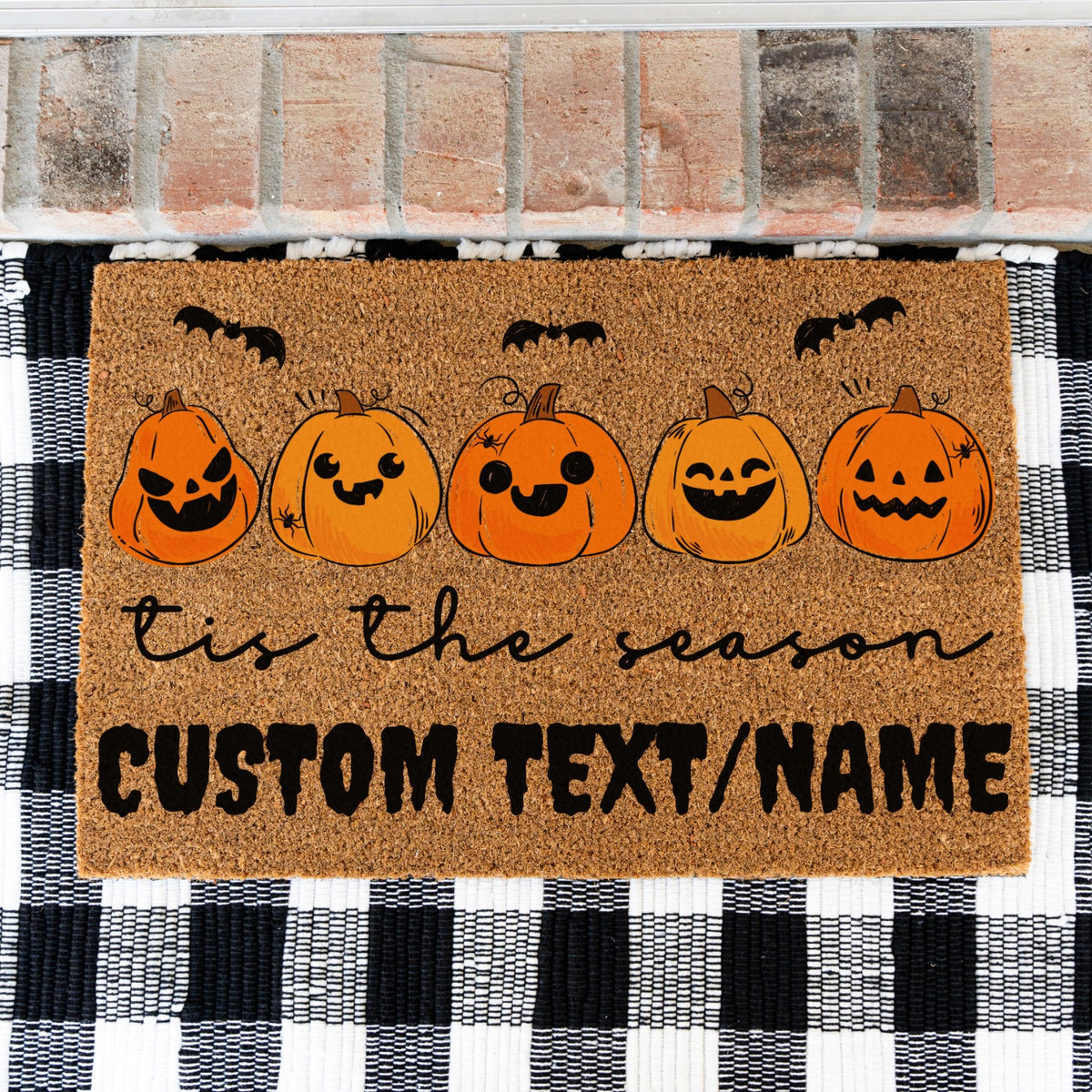 A rectangular coir doormat with a Halloween theme, featuring five smiling jack-o'-lantern pumpkins, black bats, and the text 'tis the season CUSTOM TEXT/NAME', displayed on a black and white buffalo plaid rug in front of a brick wall.