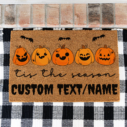 A rectangular coir doormat with a Halloween theme, featuring five smiling jack-o'-lantern pumpkins, black bats, and the text 'tis the season CUSTOM TEXT/NAME', displayed on a black and white buffalo plaid rug in front of a brick wall.