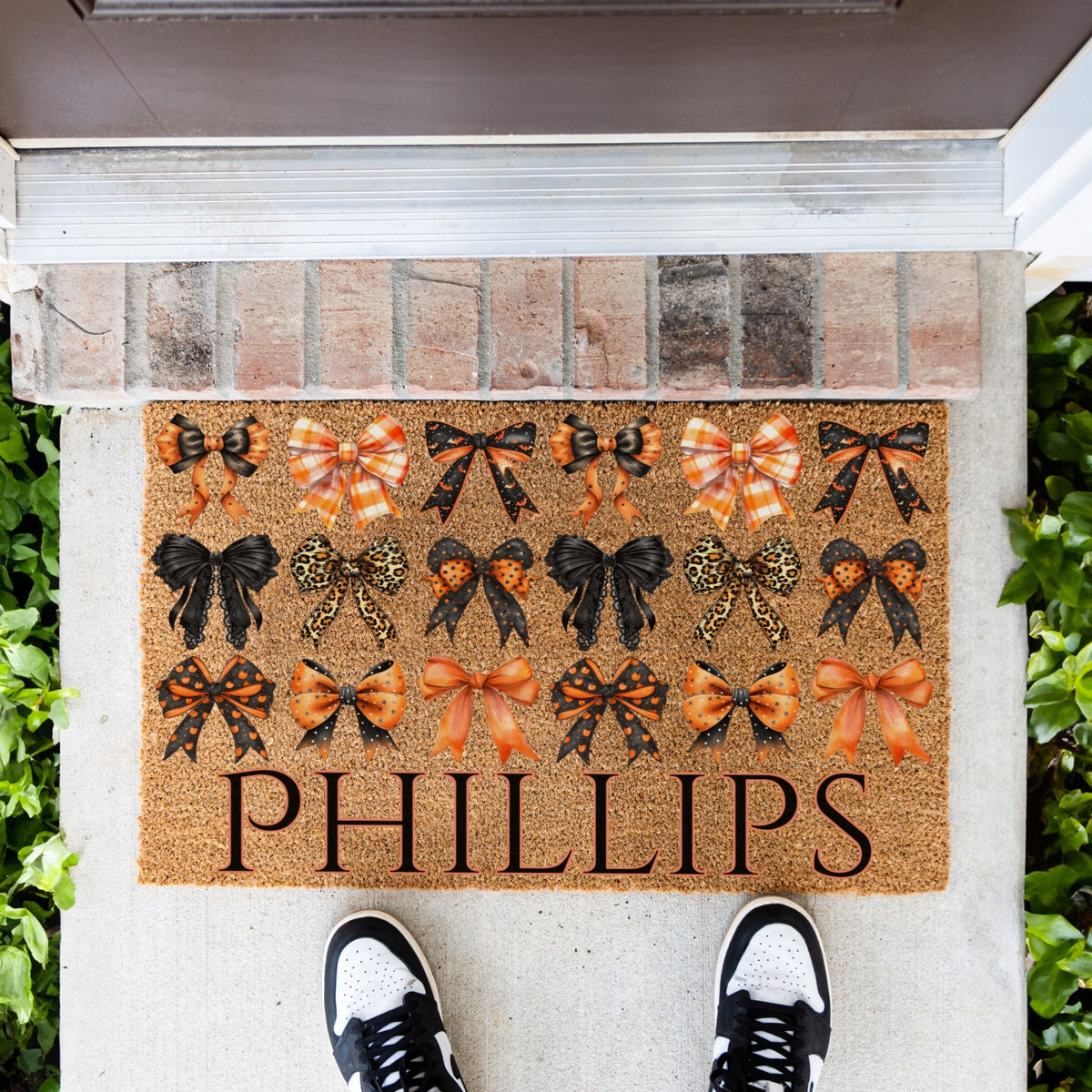 A personalized coir doormat with the custom name 'PHILLIPS' printed in large black letters, decorated with rows of black, orange, and leopard-print bow designs. The doormat is shown on a concrete porch step with a brick threshold, green plants on both sides, and a person's black and white sneakers visible at the bottom.