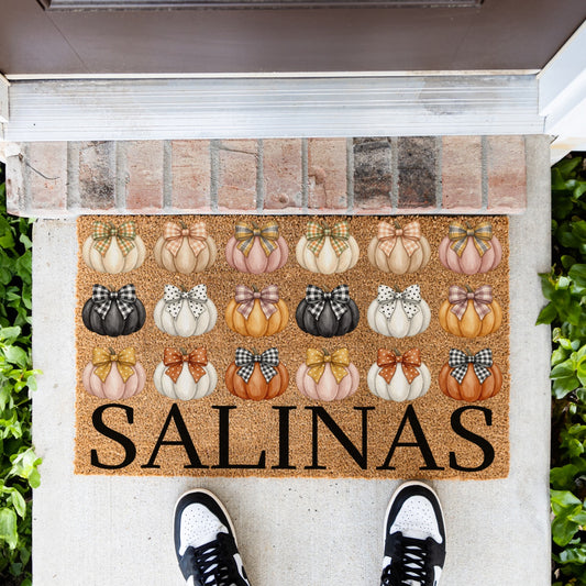 A rectangular doormat with a fall harvest theme featuring rows of illustrated pumpkins in orange, cream, black, and pink with bows, and the custom word 'SALINAS' in large black letters. The mat is placed on a concrete porch step with a brick threshold, green plants on both sides, and a person's feet in black and white sneakers standing at the bottom.