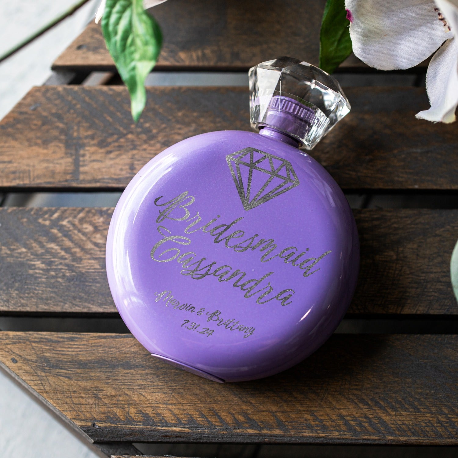 A round lavender purple flask with a clear faceted cap, personalized with 'Bridesmaid Cassandra' and a diamond graphic, resting on a dark wooden slatted surface with white flowers in the background.