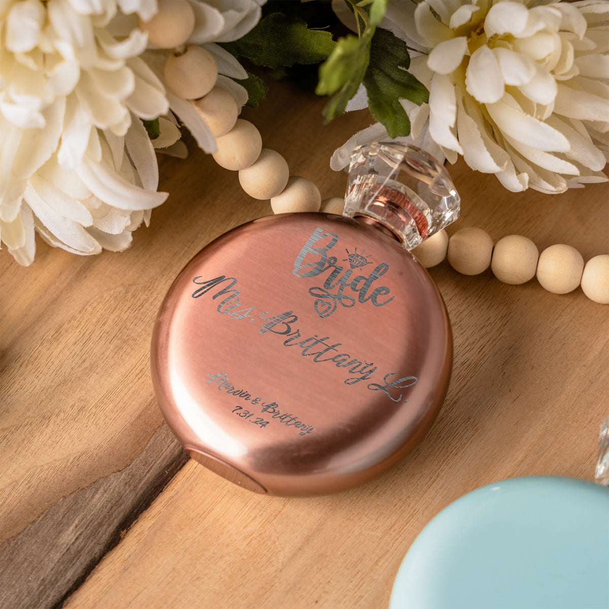 A round rose gold flask bottle engraved with 'Bride Mrs. Brittany L' in cursive, styled on a wooden surface with white flowers and wooden beads.