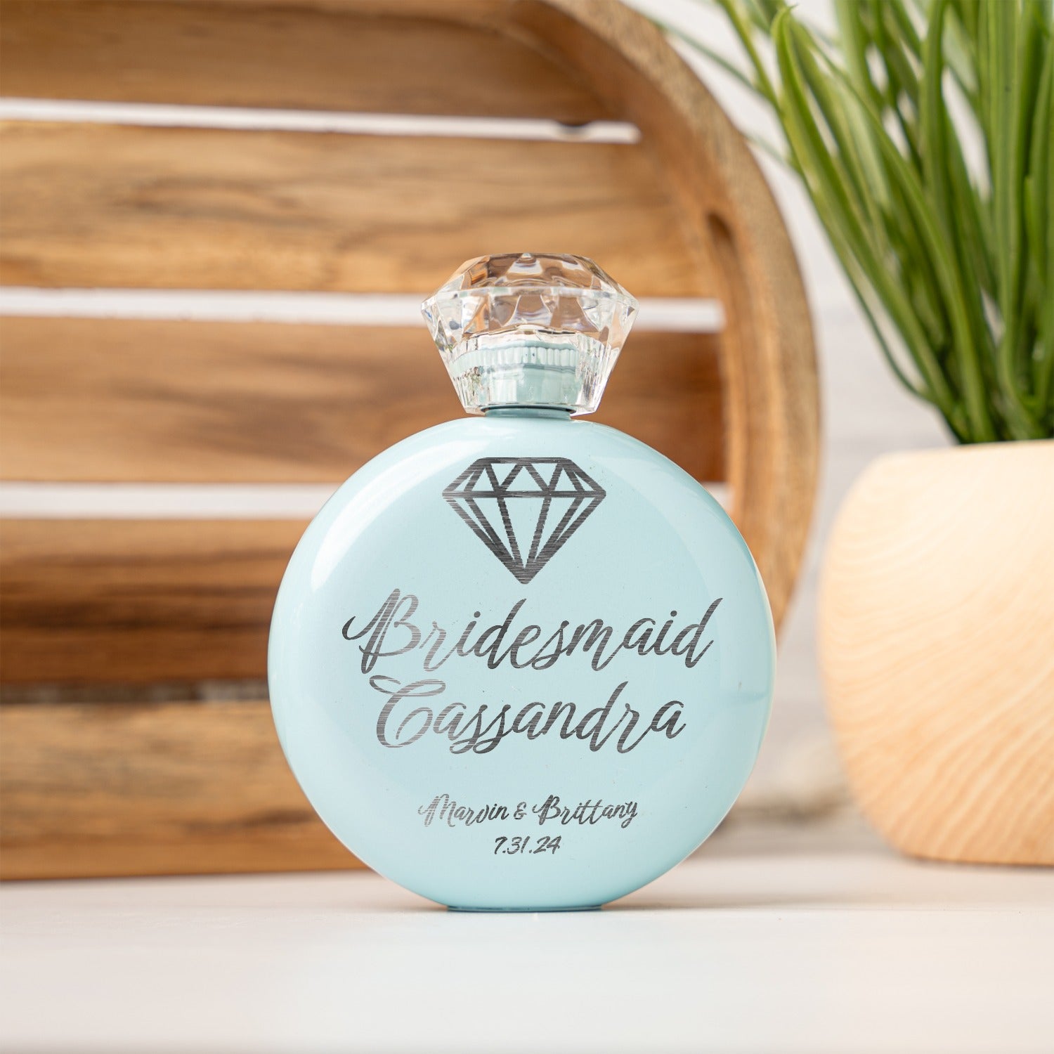 A personalized light blue round keepsake flask with a faceted clear cap, featuring a diamond graphic and the custom text 'Bridesmaid Cassandra' and 'Marvin & Brittany and the date.