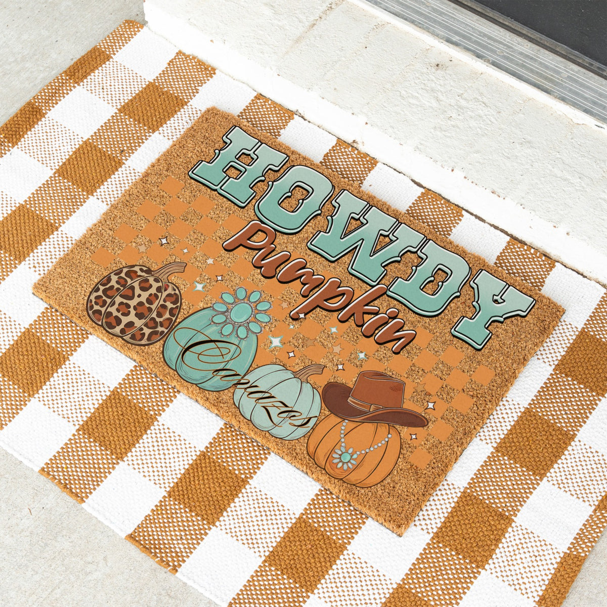 A rectangular fall-themed doormat with the words “HOWDY pumpkin” and illustrated pumpkins, placed on a white and tan gingham blanket outdoors near a door threshold.
