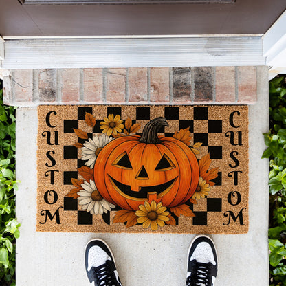 A custom Halloween doormat featuring a large orange jack-o'-lantern, black and tan checkerboard pattern, yellow and white flowers, and the word 'CUSTOM' printed vertically on both sides. The doormat is shown on a concrete porch with a brick threshold and a person's feet in black and white sneakers visible at the bottom.
