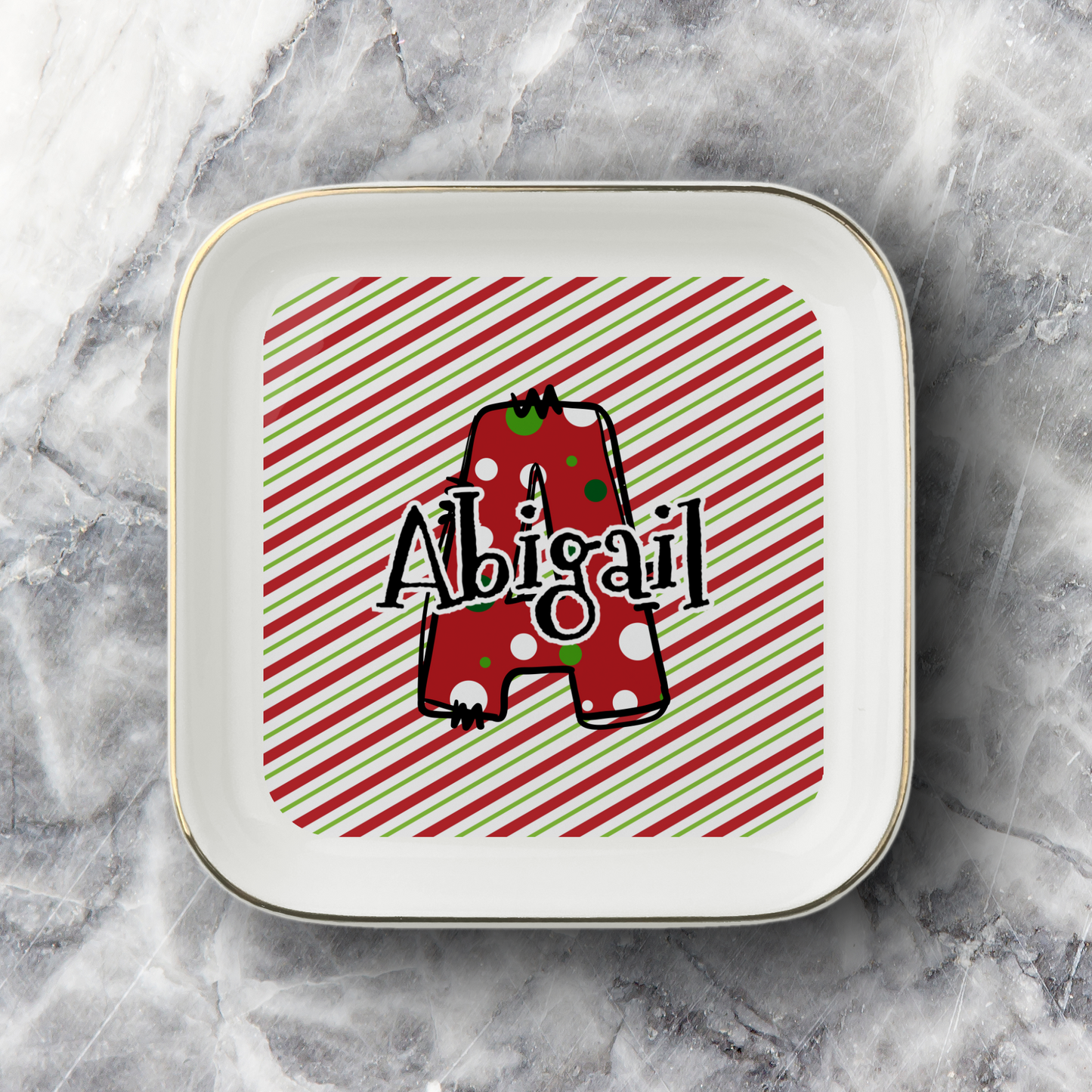 A square white ceramic jewelry dish with rounded corners and a thin gold rim, featuring a personalized design with a red and green striped background, a red polka-dot letter 'A', and the name 'Abigail' in black script. The jewelry dish is photographed on a gray and white marble surface.