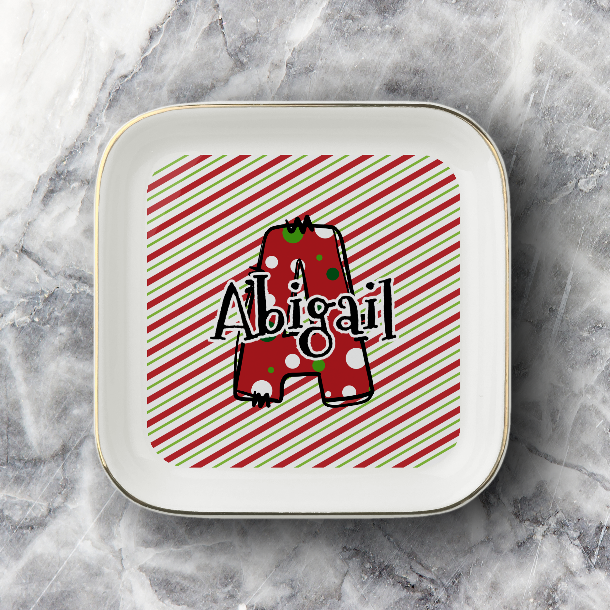 A square white ceramic jewelry dish with rounded corners and a thin gold rim, featuring a personalized design with a red and green striped background, a red polka-dot letter 'A', and the name 'Abigail' in black script. The jewelry dish is photographed on a gray and white marble surface.