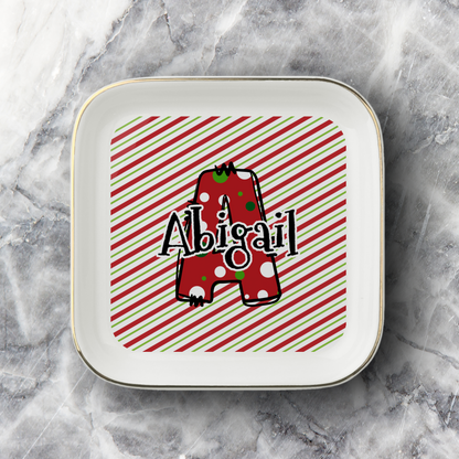 A square white ceramic jewelry dish with rounded corners and a thin gold rim, featuring a personalized design with a red and green striped background, a red polka-dot letter 'A', and the name 'Abigail' in black script. The jewelry dish is photographed on a gray and white marble surface.