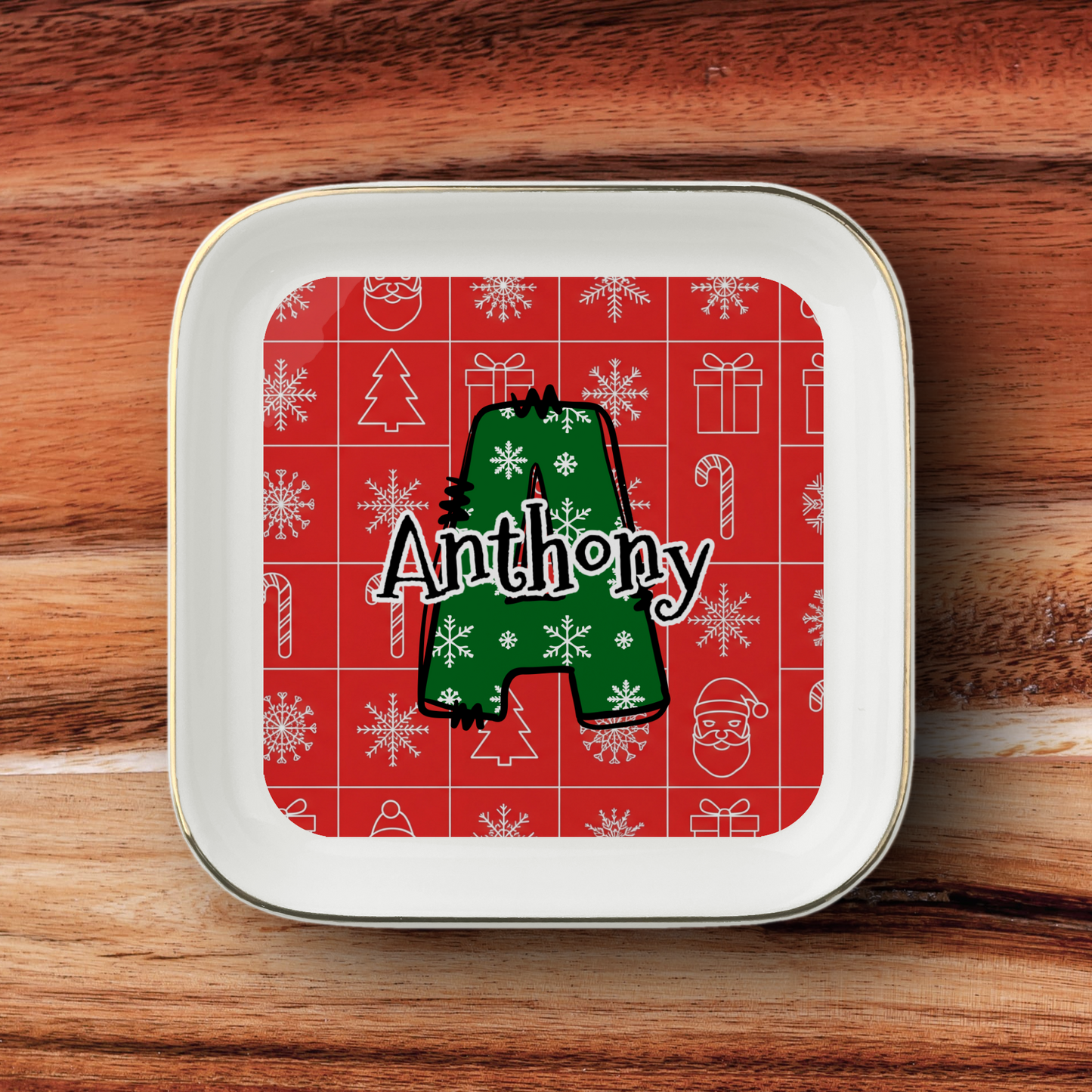 A square white ceramic tray with rounded corners and a thin gold rim, featuring a red Christmas-themed printed insert with white holiday icons and a large green letter 'A' overlaid with the name 'Anthony' in white script. The tray is photographed from above on a brown wooden surface.