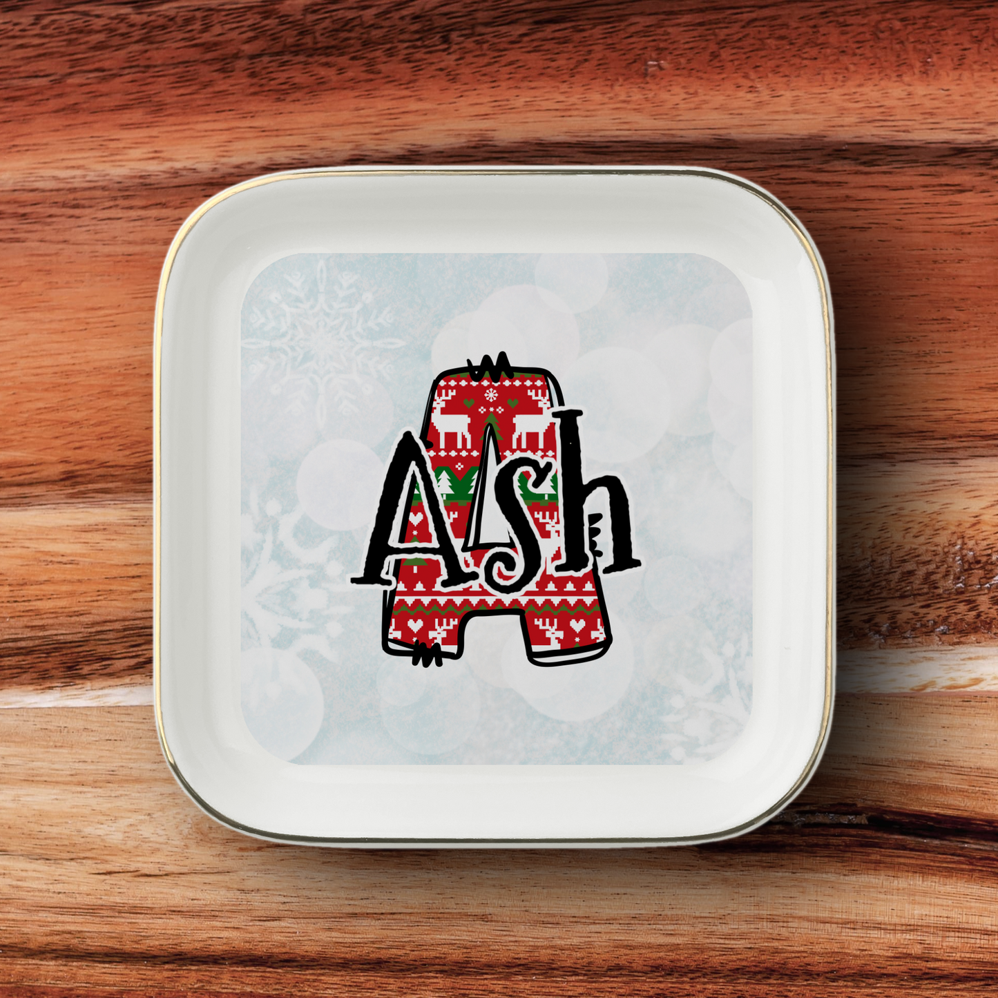 A square white ceramic plate with rounded corners and a gold rim, featuring a custom monogram design of the letter 'A' filled with a red Christmas sweater pattern and the name 'Ash' in black script. The plate is photographed flat on a warm brown wooden surface.