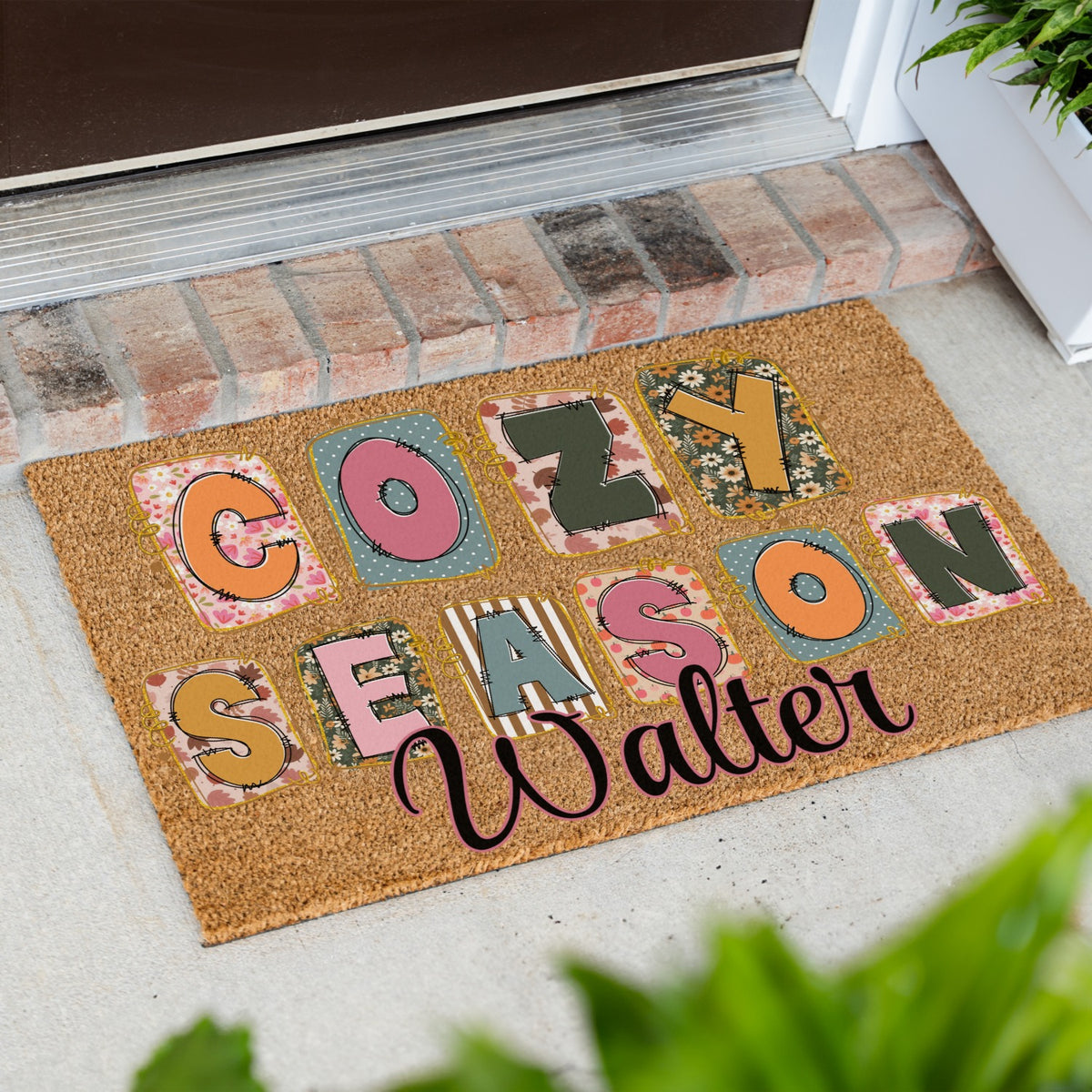 A personalized coir doormat with the words 'COZY SEASON' in colorful patterned letters and the custom name 'Walter' in black script, placed at a front door on a concrete step.