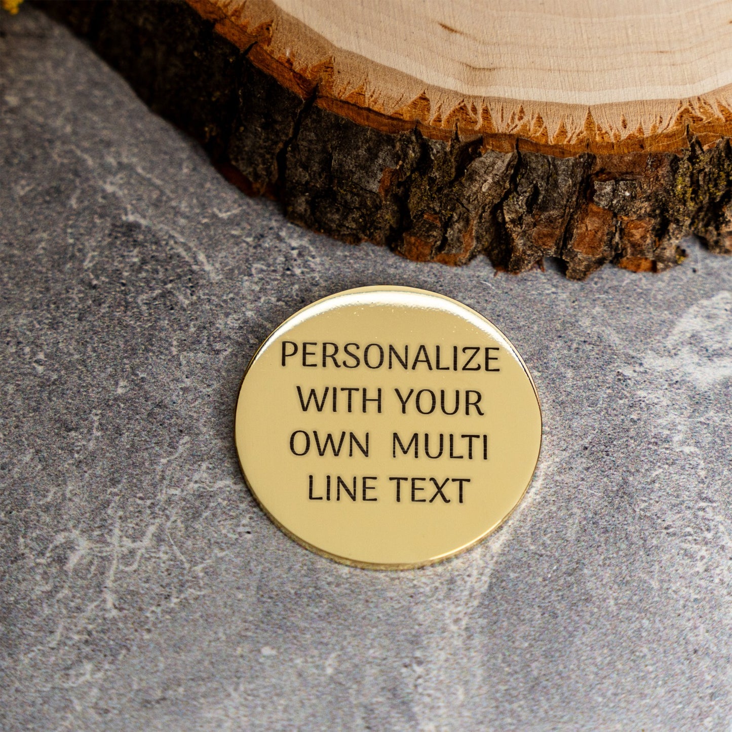 A round, gold-toned metal coin engraved with the custom text 'PERSONALIZE WITH YOUR OWN MULTI LINE TEXT' in black lettering, resting on a textured gray stone surface next to a rustic wood slice.