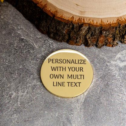 A round, gold-toned metal coin engraved with the custom text 'PERSONALIZE WITH YOUR OWN MULTI LINE TEXT' in black lettering, resting on a textured gray stone surface next to a rustic wood slice.