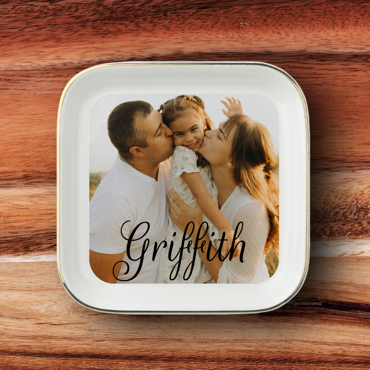 A square white ceramic plate with rounded corners and a thin metallic rim, featuring a custom printed photo of a family of three and the name 'Griffith' in black script. The plate is placed on a rustic wooden surface.