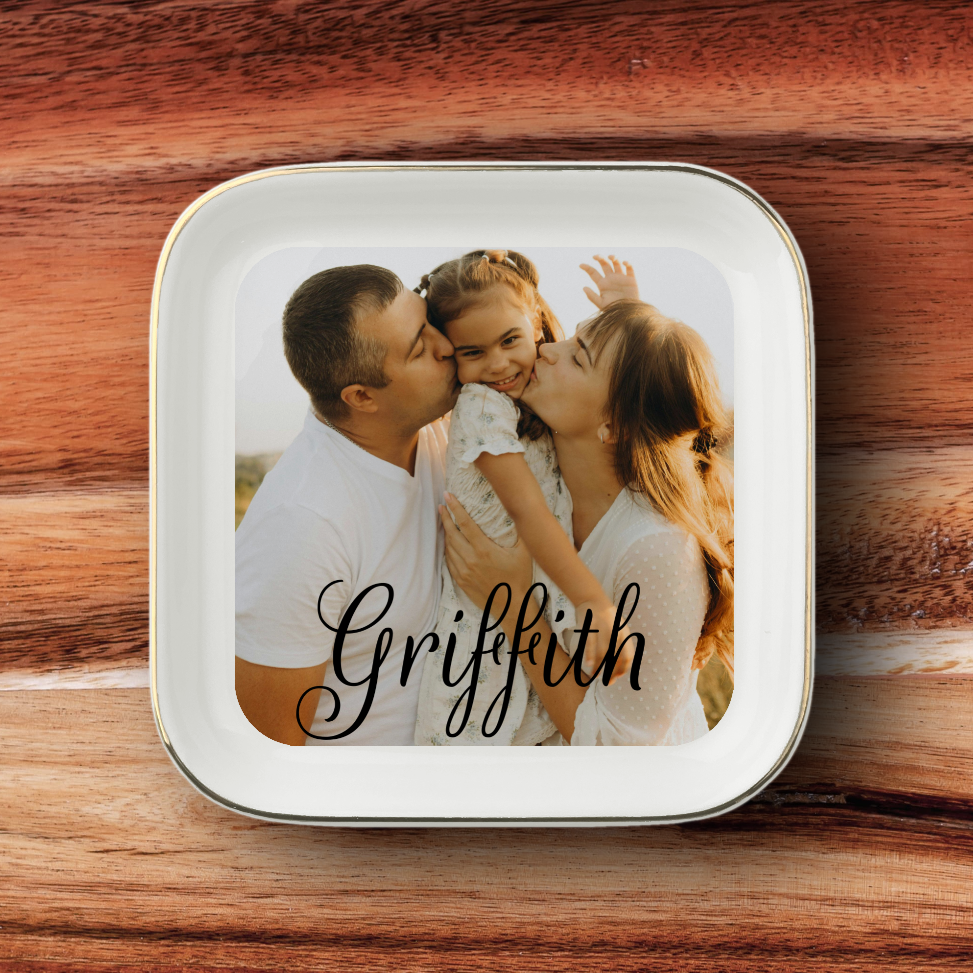 A square white ceramic plate with rounded corners and a thin metallic rim, featuring a custom printed photo of a family of three and the name 'Griffith' in black script. The plate is placed on a rustic wooden surface.