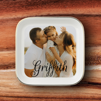 A square white ceramic plate with rounded corners and a thin metallic rim, featuring a custom printed photo of a family of three and the name 'Griffith' in black script. The plate is placed on a rustic wooden surface.