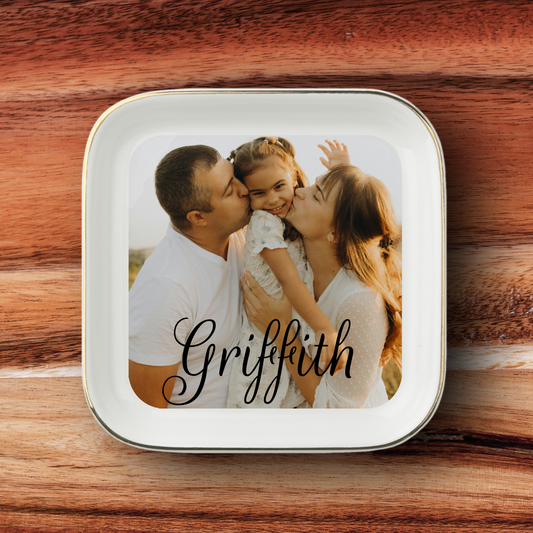 A square white ceramic plate with rounded corners and a thin metallic rim, featuring a custom printed photo of a family of three and the name 'Griffith' in black script. The plate is placed on a rustic wooden surface.