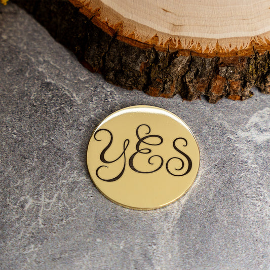 A round gold-toned coin with the custom word 'YES' in black cursive script, placed on a gray stone surface with a rustic wood slice in the background.