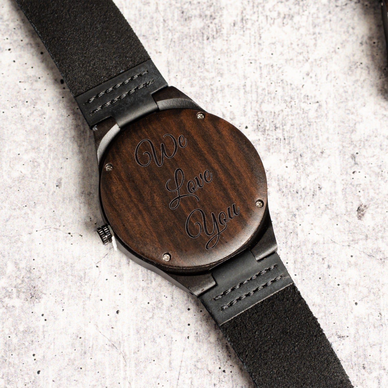 A close-up of a wristwatch shown from the back, featuring a round dark wood case custom engraved with 'We Love You' in cursive script, paired with a black textured strap, set against a light gray concrete background.