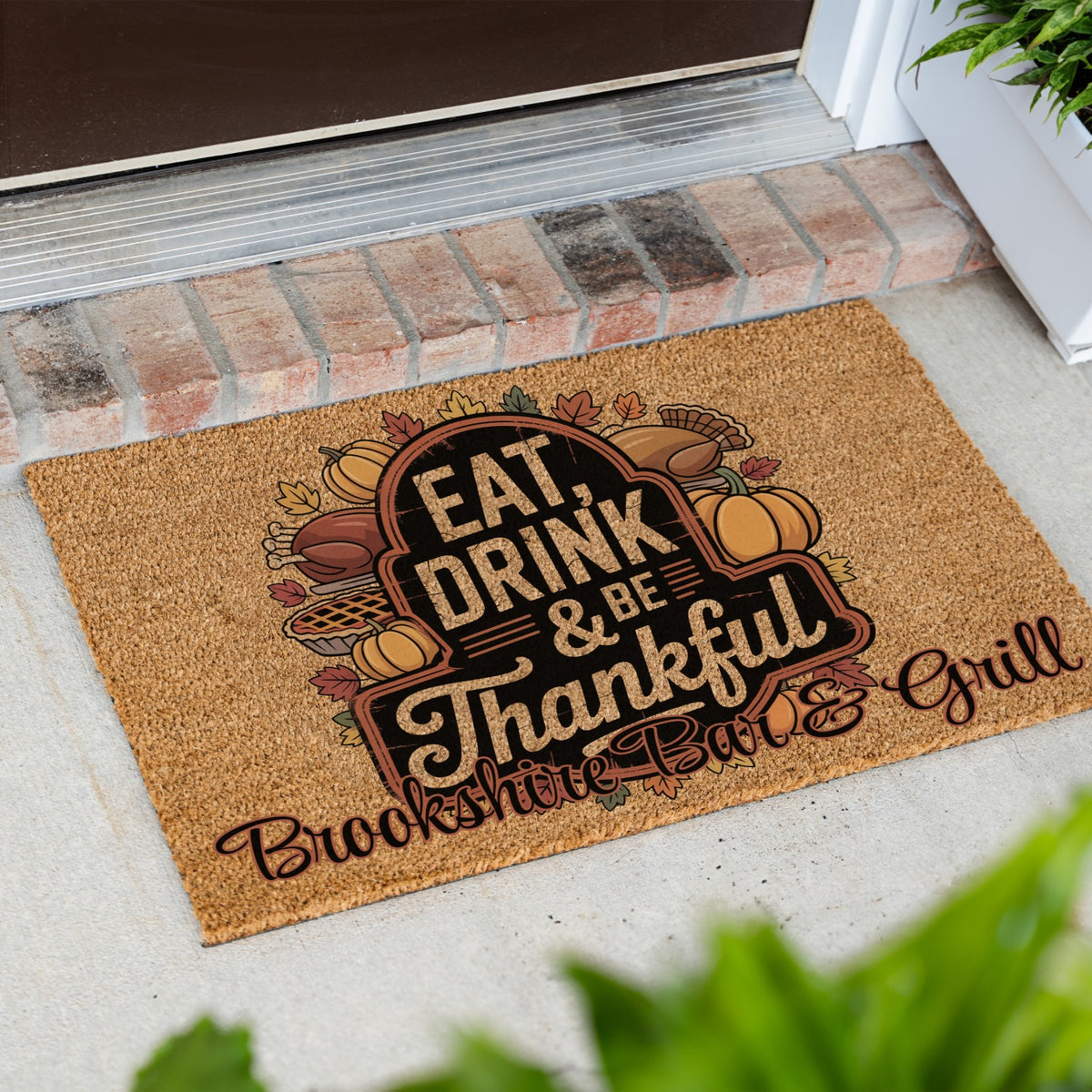 A rectangular coir doormat with a fall-themed graphic reading 'EAT, DRINK & BE Thankful' and 'Brookshire Bar & Grill,' placed on a concrete porch step with brick trim and greenery visible.