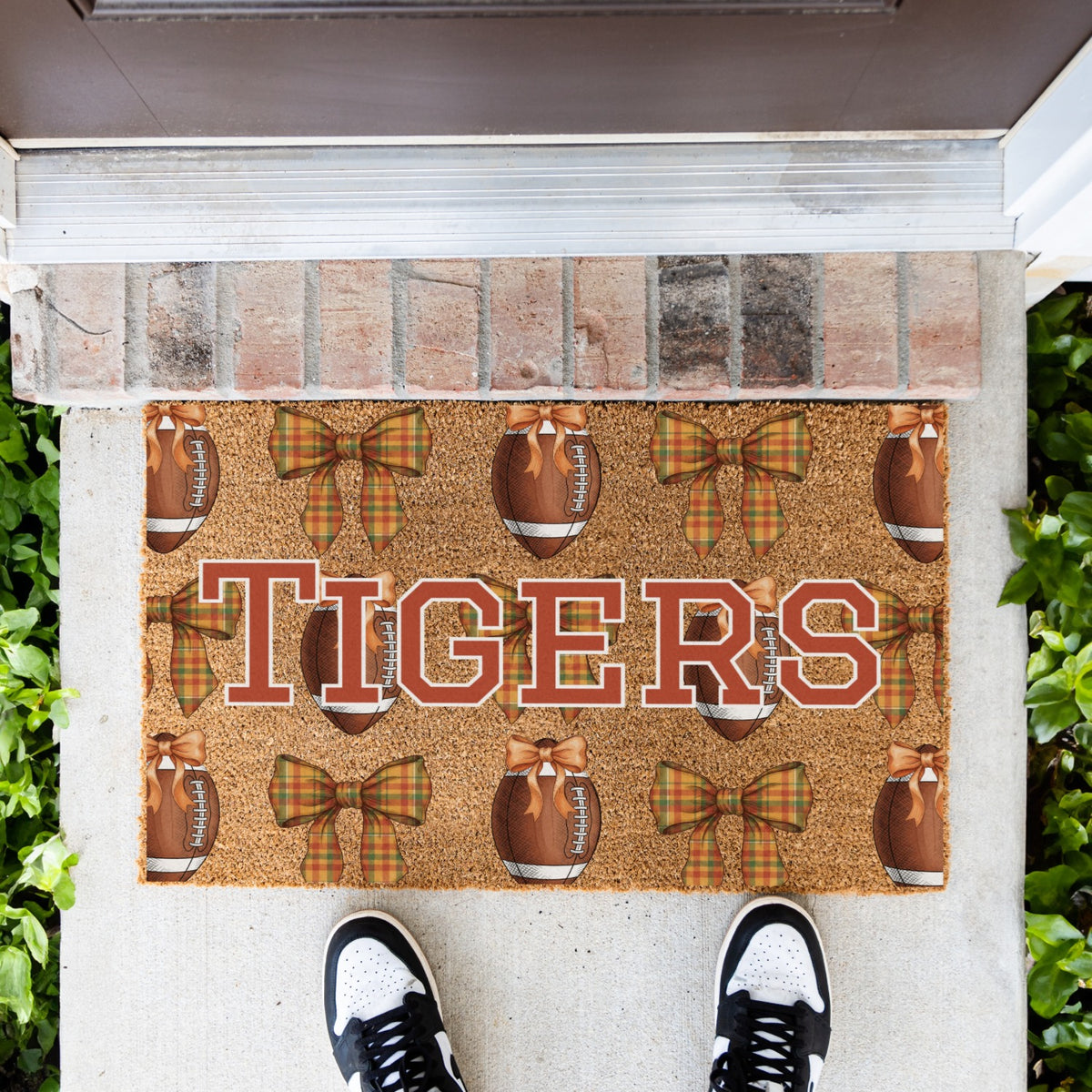 A rectangular coir doormat with a brown and orange custom word 'TIGERS' text design, footballs, and plaid bow ties, shown on a concrete porch with a brick threshold and a person's shoes visible at the bottom.