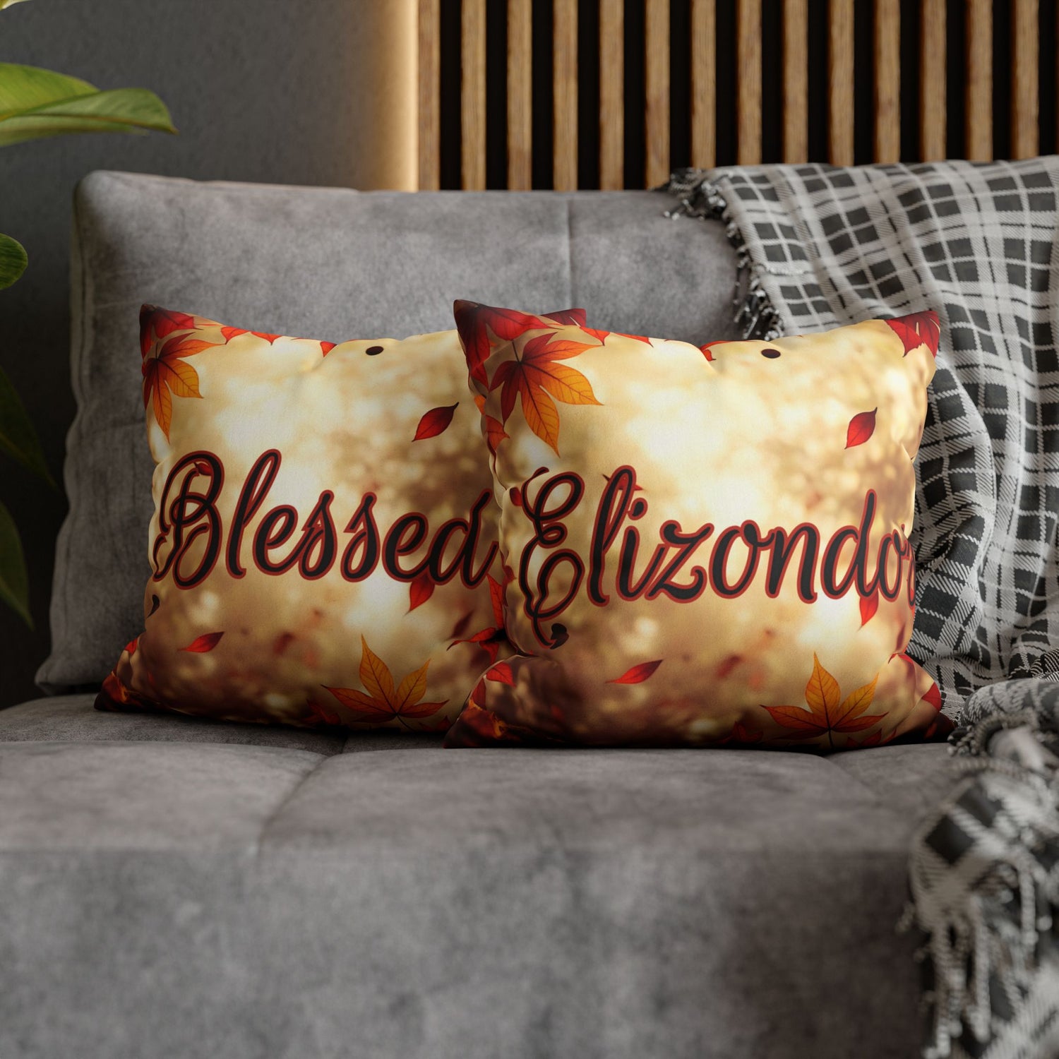 Two decorative throw pillows on a gray sofa, featuring a warm cream background with autumn leaf motifs and the words Blessed on one side and  Elizondo on the other in dark script. A plaid throw blanket and wooden slat wall are visible in the background.