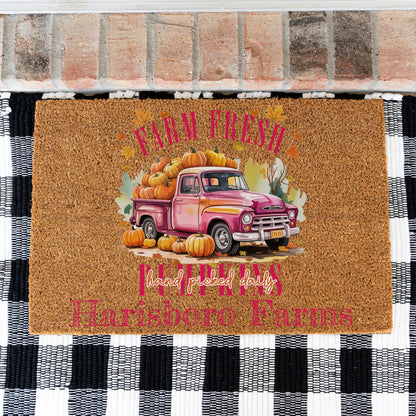 A rectangular coir doormat with a fall harvest design, placed on a black and white checkered rug in front of a brick threshold. The mat features a pink pickup truck loaded with pumpkins and the text 'FARM FRESH', 'hand picked daily', and custom text 'Harisboro Farms' on the bottom.