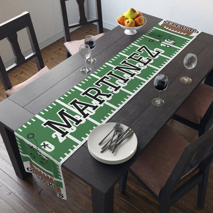 A green and white football field themed table runner with the custom name 'MARTINEZ' in large black letters, styled on a dark wooden dining table with plates, wine glasses, and chairs.