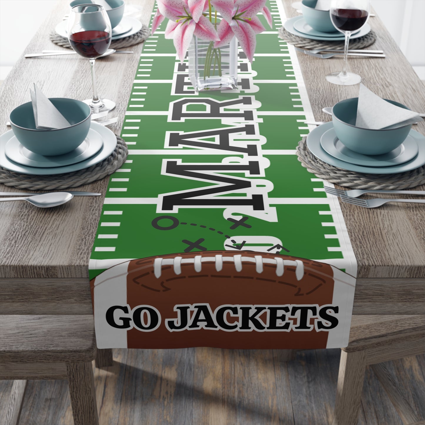 A football-themed table runner displayed on a dining table, featuring a green football field design with white yard lines, large custom 'MARTINEZ' lettering, a brown football graphic at the bottom with custom 'GO JACKETS' text