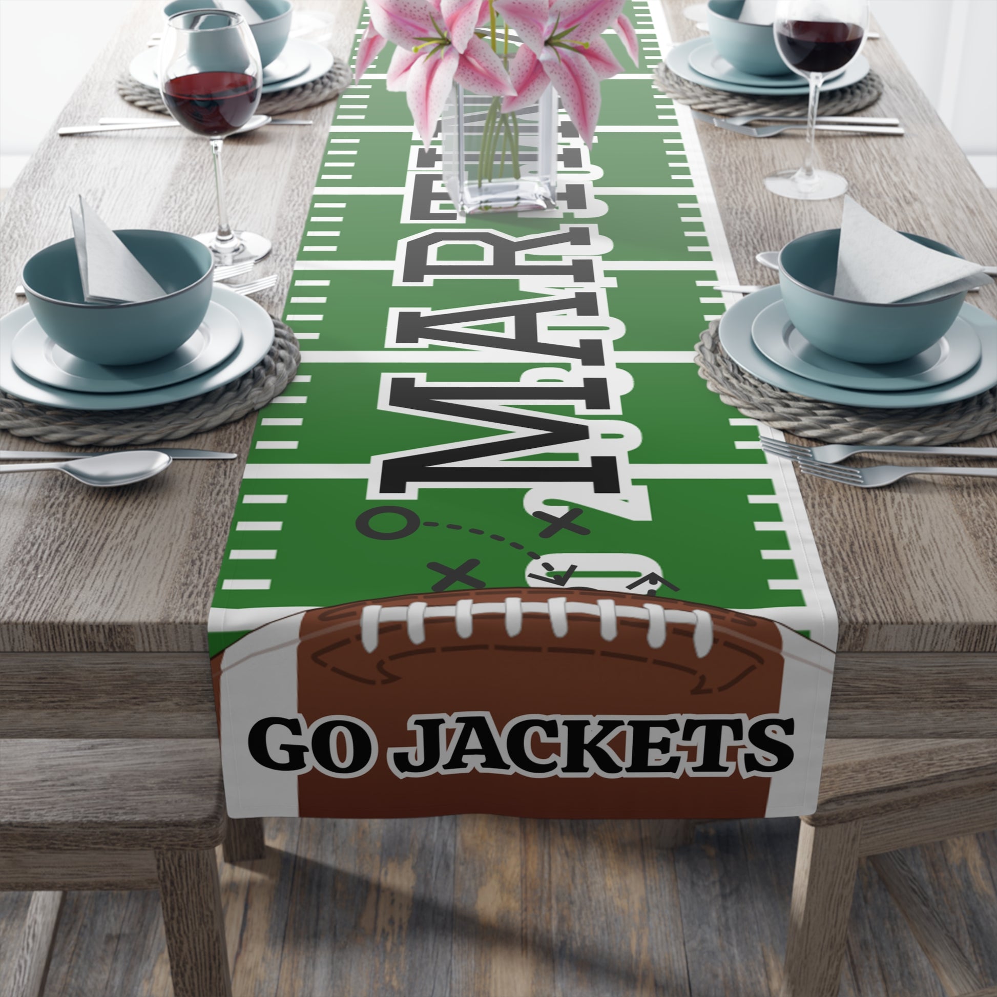 A football-themed table runner displayed on a dining table, featuring a green football field design with white yard lines, large custom 'MARTINEZ' lettering, a brown football graphic at the bottom with custom 'GO JACKETS' text
