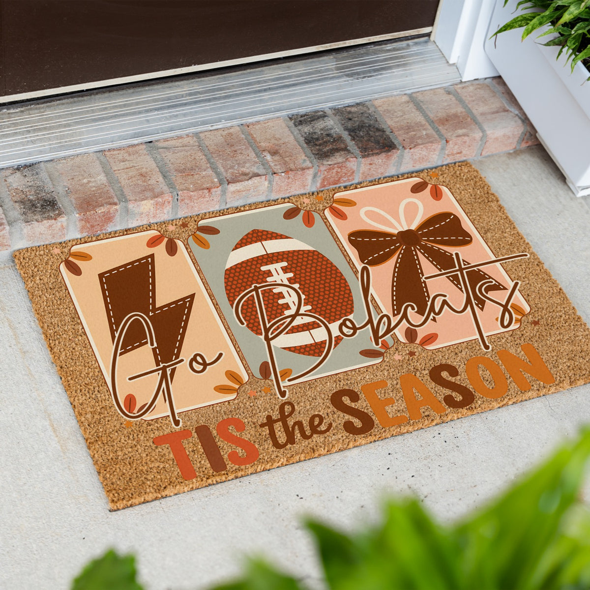 A rectangular coir doormat with a Thanksgiving football theme, placed on a concrete porch in front of a brick threshold. The mat features three panels with a lightning bolt, a football, and a bow, surrounded by autumn leaves, with the text 'Go Bobcats TIS the SEASON'.