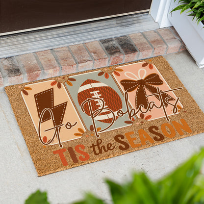 A rectangular coir doormat with a Thanksgiving football theme, placed on a concrete porch in front of a brick threshold. The mat features three panels with a lightning bolt, a football, and a bow, surrounded by autumn leaves, with the text 'Go Bobcats TIS the SEASON'.