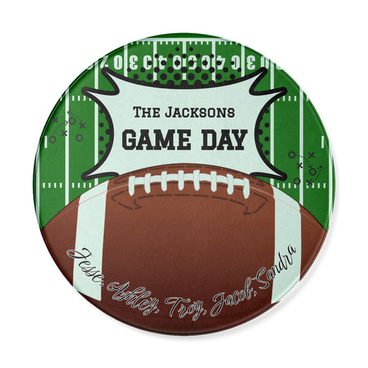 A round custom football-themed cutting board featuring a brown football on a green field background with white yard lines. A white banner across the top reads 'THE JACKSONS GAME DAY' in black text, and the bottom shows custom names cursive text.