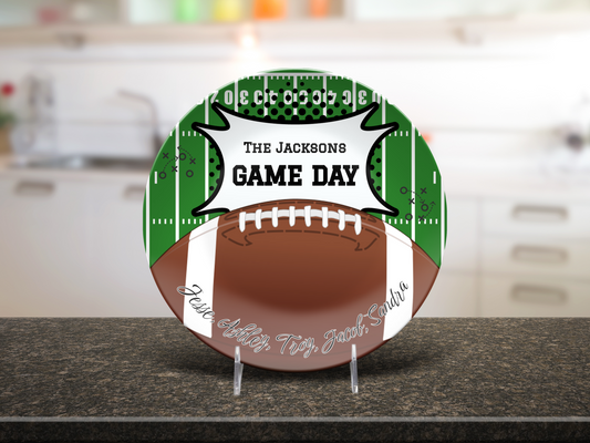 A round decorative plate displayed on clear stands on a dark countertop, featuring a football design with green field graphics, white laces, a brown football body, and the custom text 'THE JACKSONS GAME DAY' with custom names cursive text at the bottom.