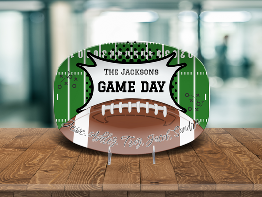 A personalized football-themed platter displayed on a wooden table. The sign features a green football field background with white yard lines, a brown football graphic in the center, and a white banner reading custom tex 'THE JACKSONS GAME DAY'. and custom names cursive text on the bottom