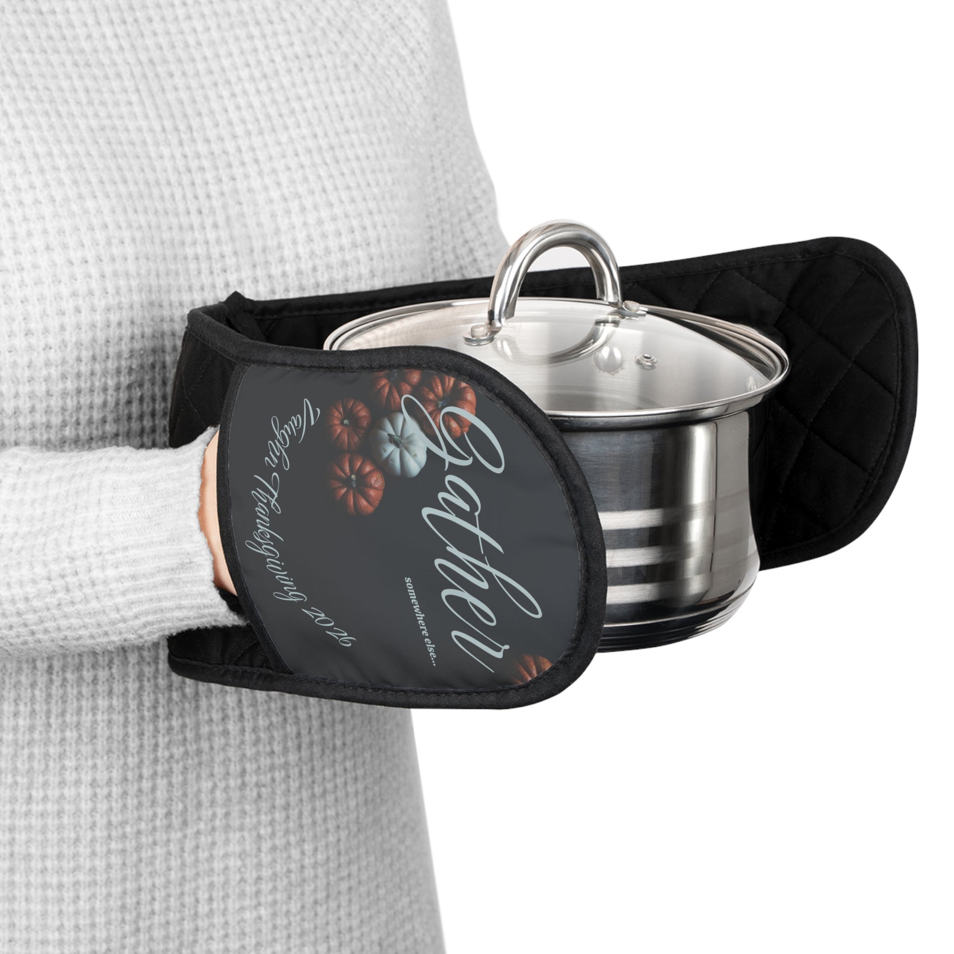 Person holding a pot with a patterned oven mitt on a white background. The oven mit is custom text on the bottom and reads Gather somewhere else with some pumpkins behind the words