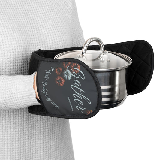 Person holding a pot with a patterned oven mitt on a white background. The oven mit is custom text on the bottom and reads Gather somewhere else with some pumpkins behind the words