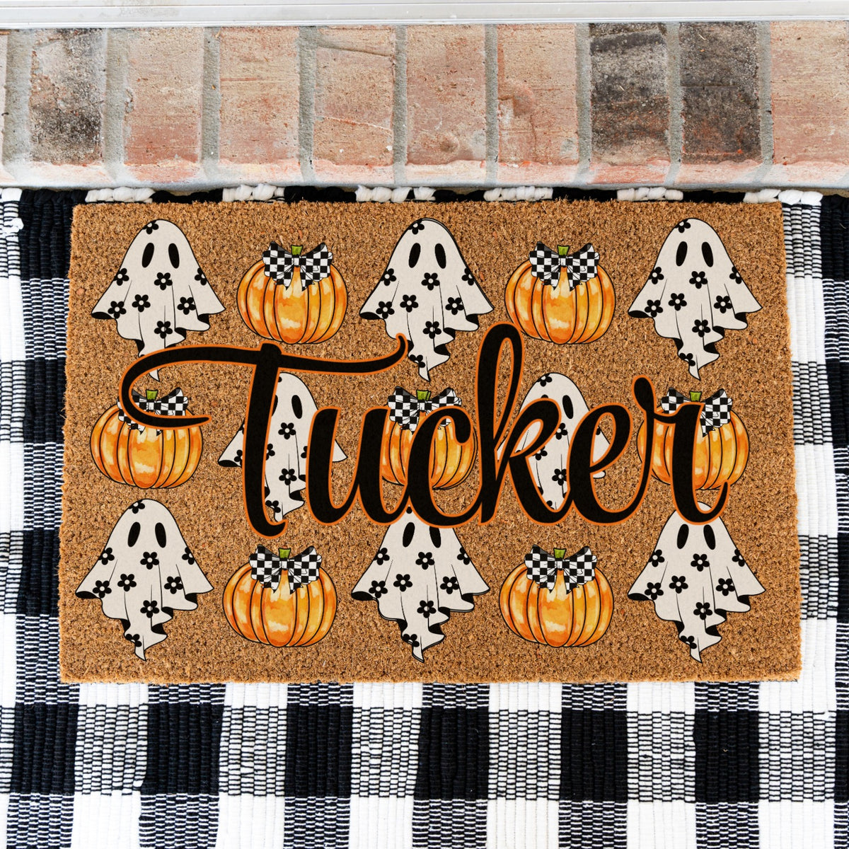 A rectangular coir doormat with a Halloween theme, featuring the name 'Tucker' in large black script, orange pumpkins, and white ghosts with black flowers. The doormat is placed on a black and white checkered rug in front of a brick wall.