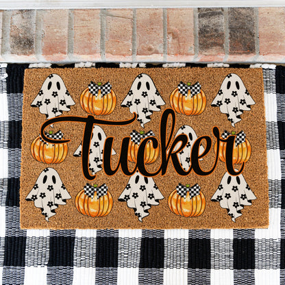 A rectangular coir doormat with a Halloween theme, featuring the name 'Tucker' in large black script, orange pumpkins, and white ghosts with black flowers. The doormat is placed on a black and white checkered rug in front of a brick wall.