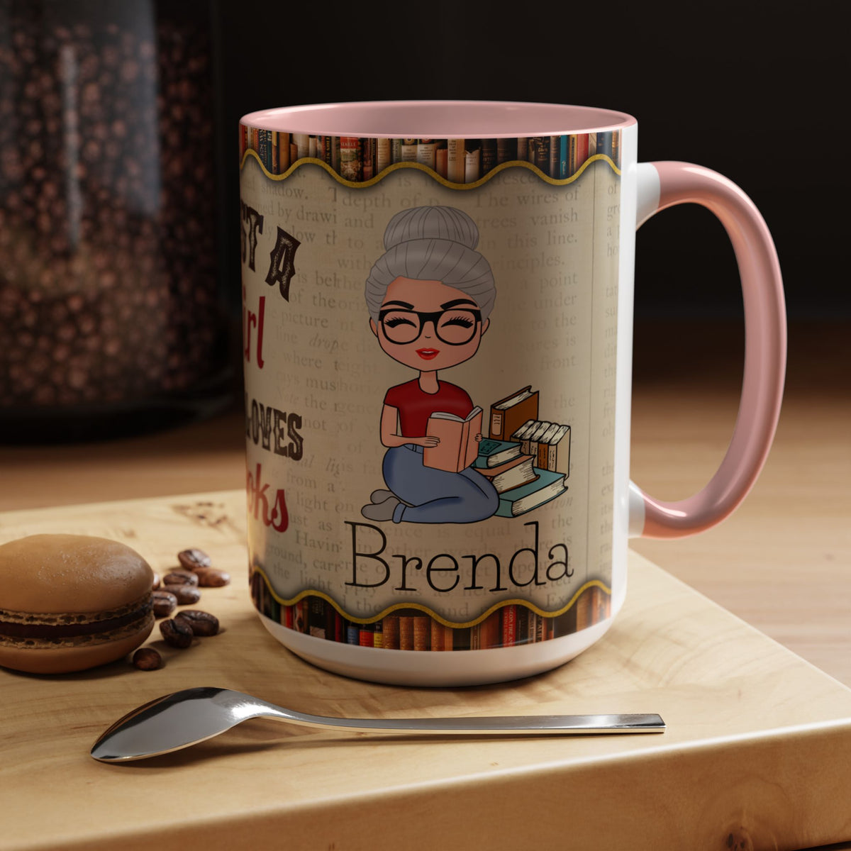 A personalized ceramic mug with a pink handle and interior, featuring a cartoon illustration of a woman reading and the name 'Brenda' printed on a light wood board with a spoon and macaroni nearby.