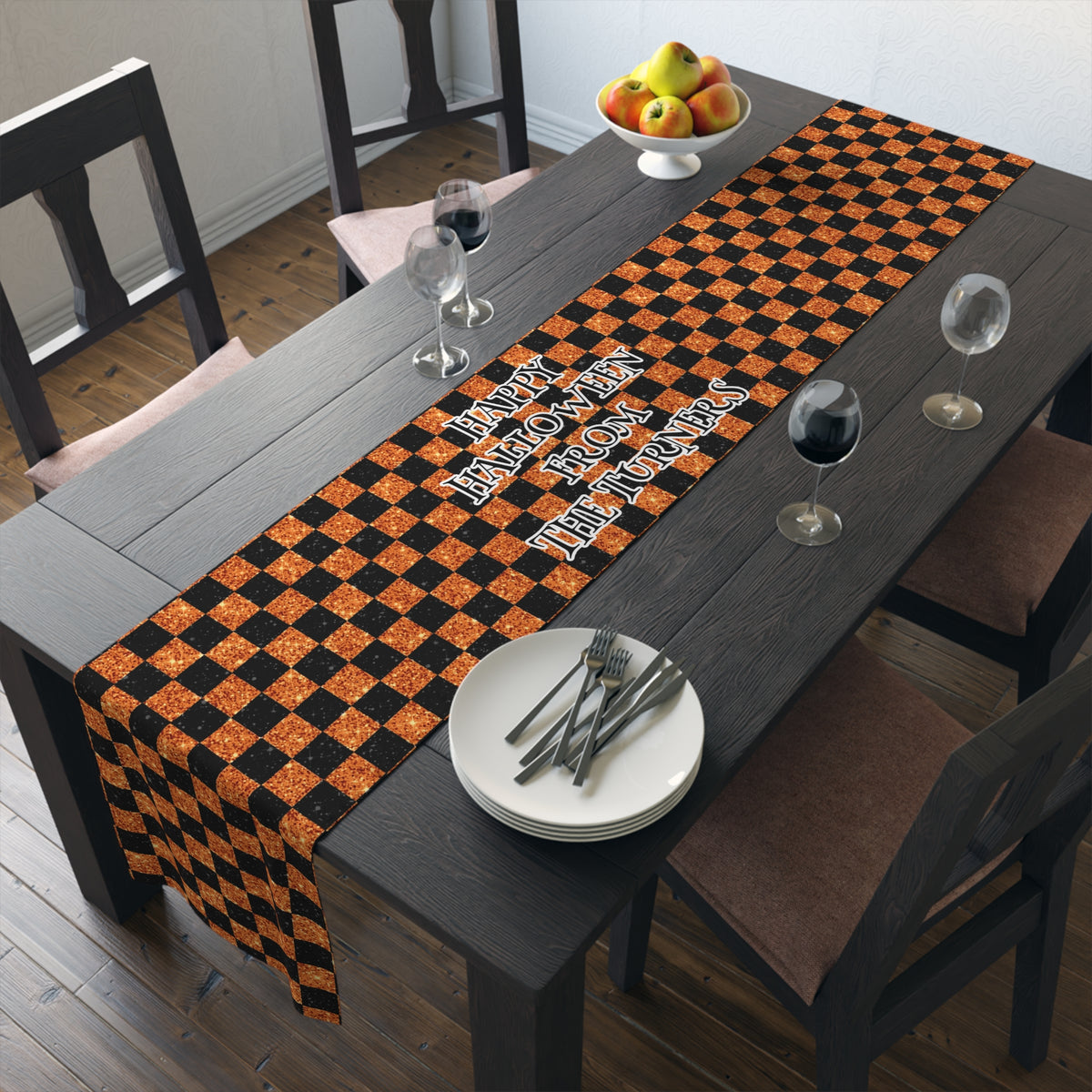 A rectangular table runner with a black and orange checkered pattern and the custom text 'Happy Halloween From The Turners' is displayed on a dark wooden dining table. The table is styled with white plates, silver cutlery, wine glasses, and a bowl of fruit, surrounded by dark wooden chairs.