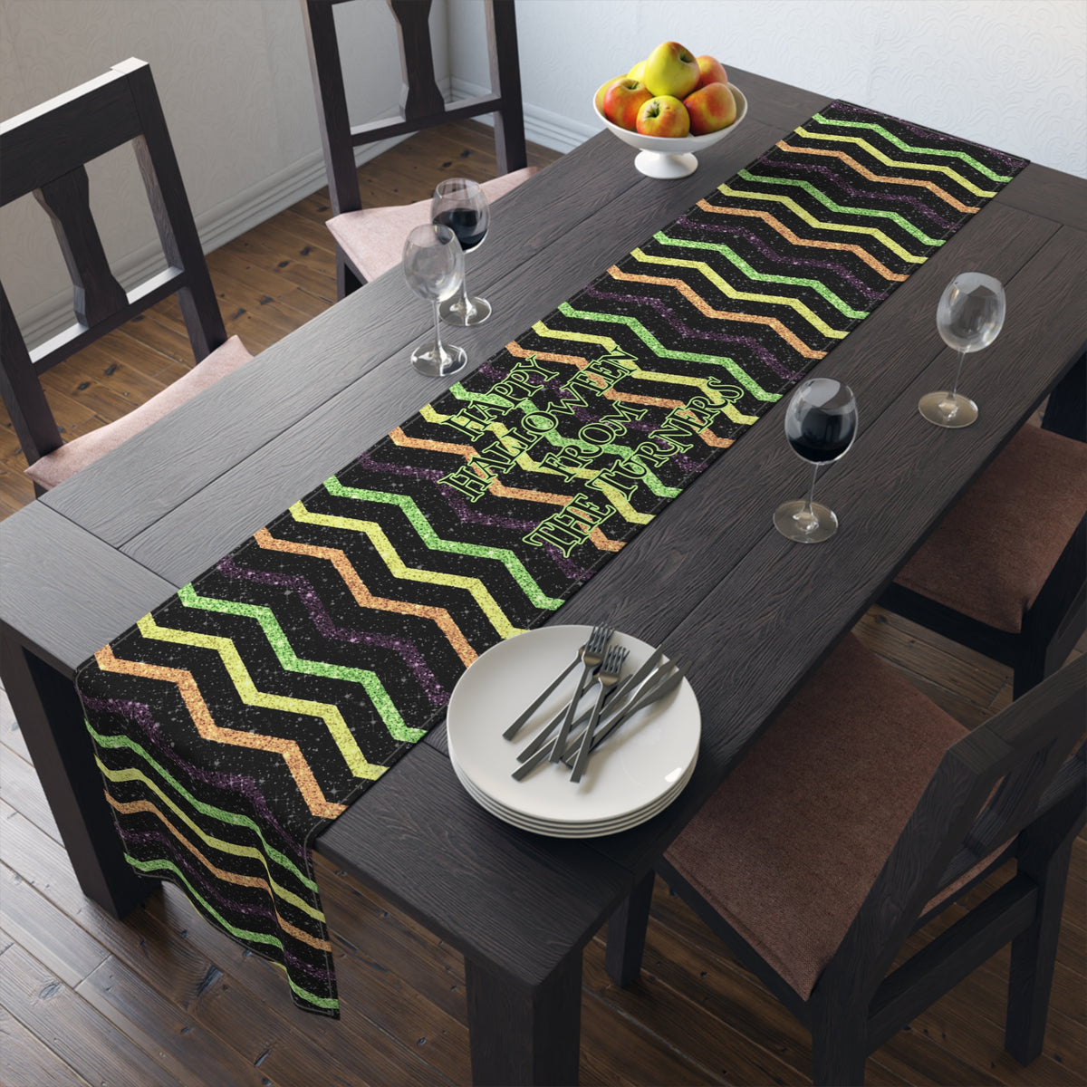 Dining table with a halloween colored zigzag / chevron pattern that looks glittery but its not table runner that has custom wording that reads HAPPY HALLOWEEN FROM THE TURNERS. There are plates, and glasses on a brown table.