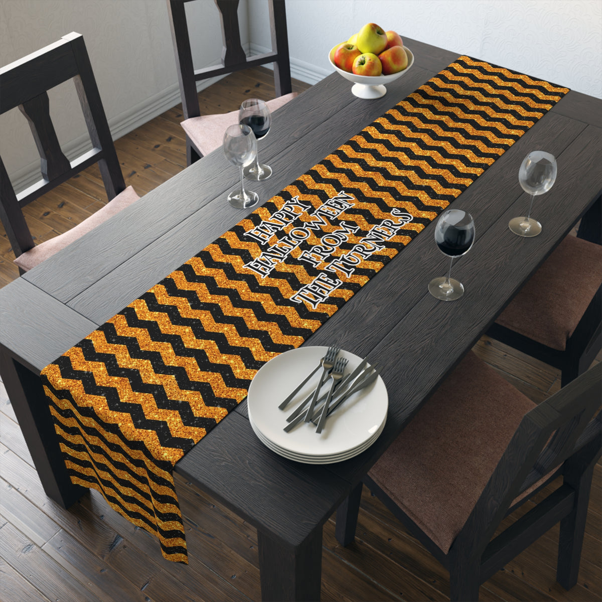 A black and orange glitter look but its not glitter chevron table runner with the custom text 'Happy Halloween From The Turners' laid out on a dark wooden dining table with plates, cutlery, wine glasses, and chairs.