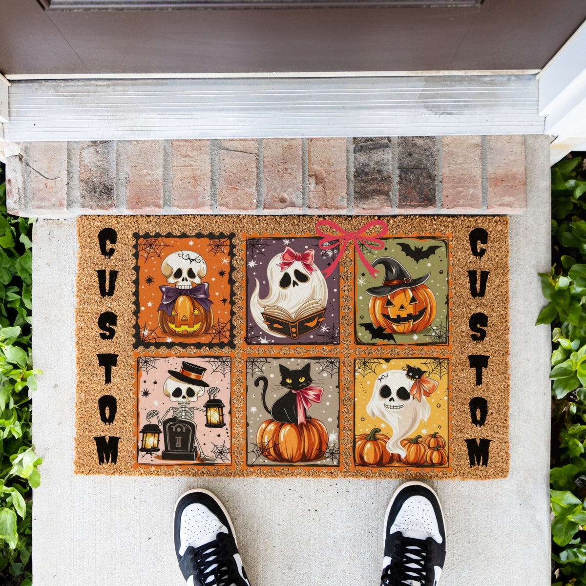 A custom Halloween doormat featuring six illustrated panels with pumpkins, ghosts, a black cat, bats, and lanterns, bordered by the word 'CUSTOM' on the left and right. The mat is shown on a concrete porch with a brick threshold, green plants on both sides, and a person's feet in black and white sneakers standing at the bottom.