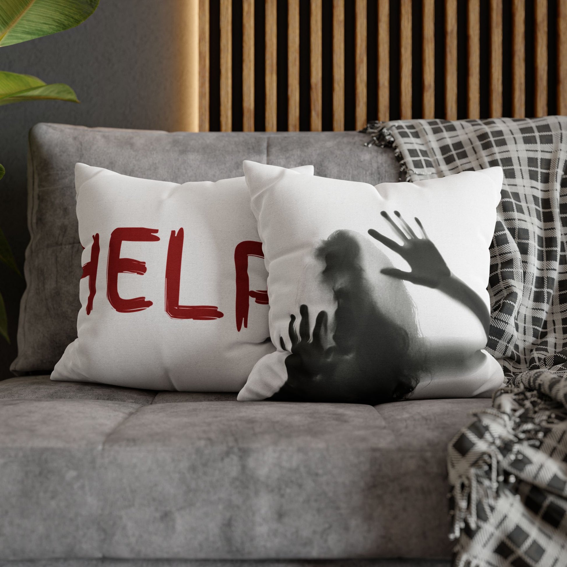 Two square throw pillows on a gray sofa in a styled living room setting. One pillow is white with red distressed lettering reading 'HELP', and the other is white with a black-and-gray distressed silhouette of a person reaching upward.