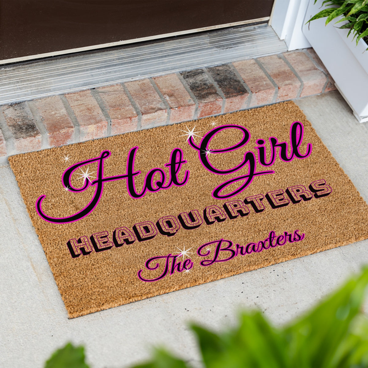 A doormat placed at a doorway, printed with the phrase “Hot Girl Headquarters The Braxters” in pink and black lettering, with decorative sparkles.