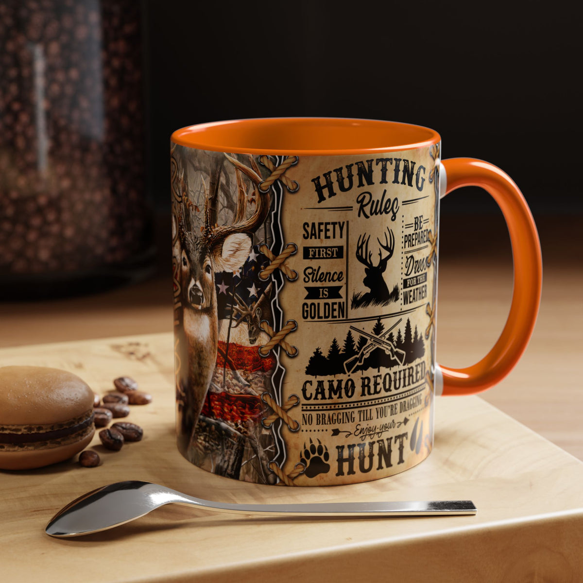 A ceramic mug with an orange handle and interior, featuring a detailed hunting-themed graphic print with deer, camouflage, and text like 'Hunting Rules' and 'Safety First'. The mug is set on a wooden surface with a spoon and coffee beans nearby.