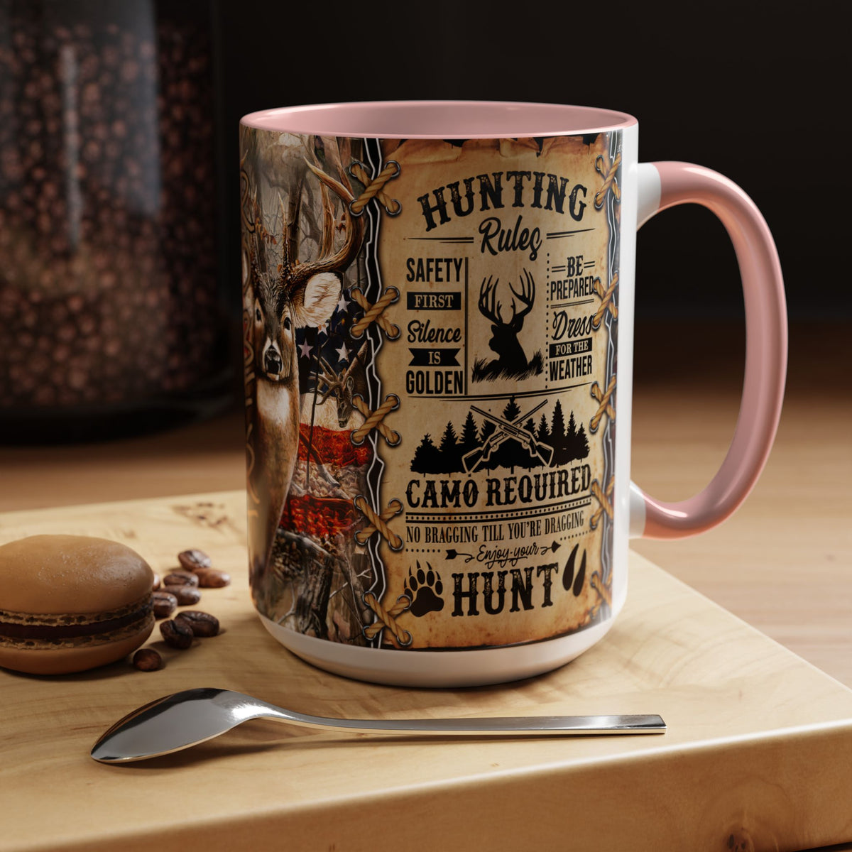 A ceramic mug with a pink handle and interior, featuring a rustic hunting-themed graphic print with text and illustrations, displayed on a wooden board with a spoon and coffee beans.