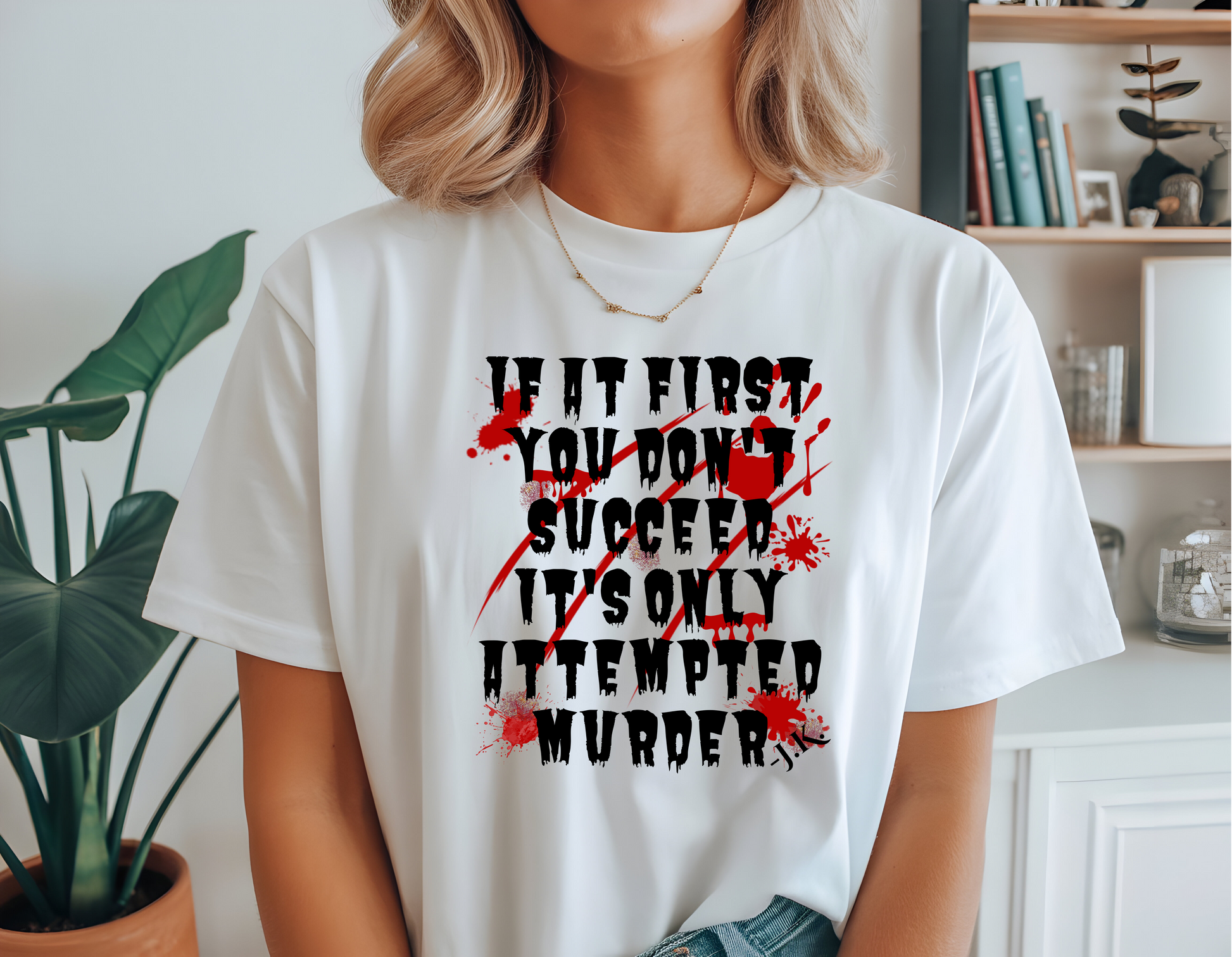 A woman wearing a white graphic t-shirt with a black and red horror-themed print that reads 'IF IT FIRST YOU DON'T SUCCEED IT'S ONLY ATTEMPTED MURDER'. The background shows a cozy indoor setting with a bookshelf and a potted plant.