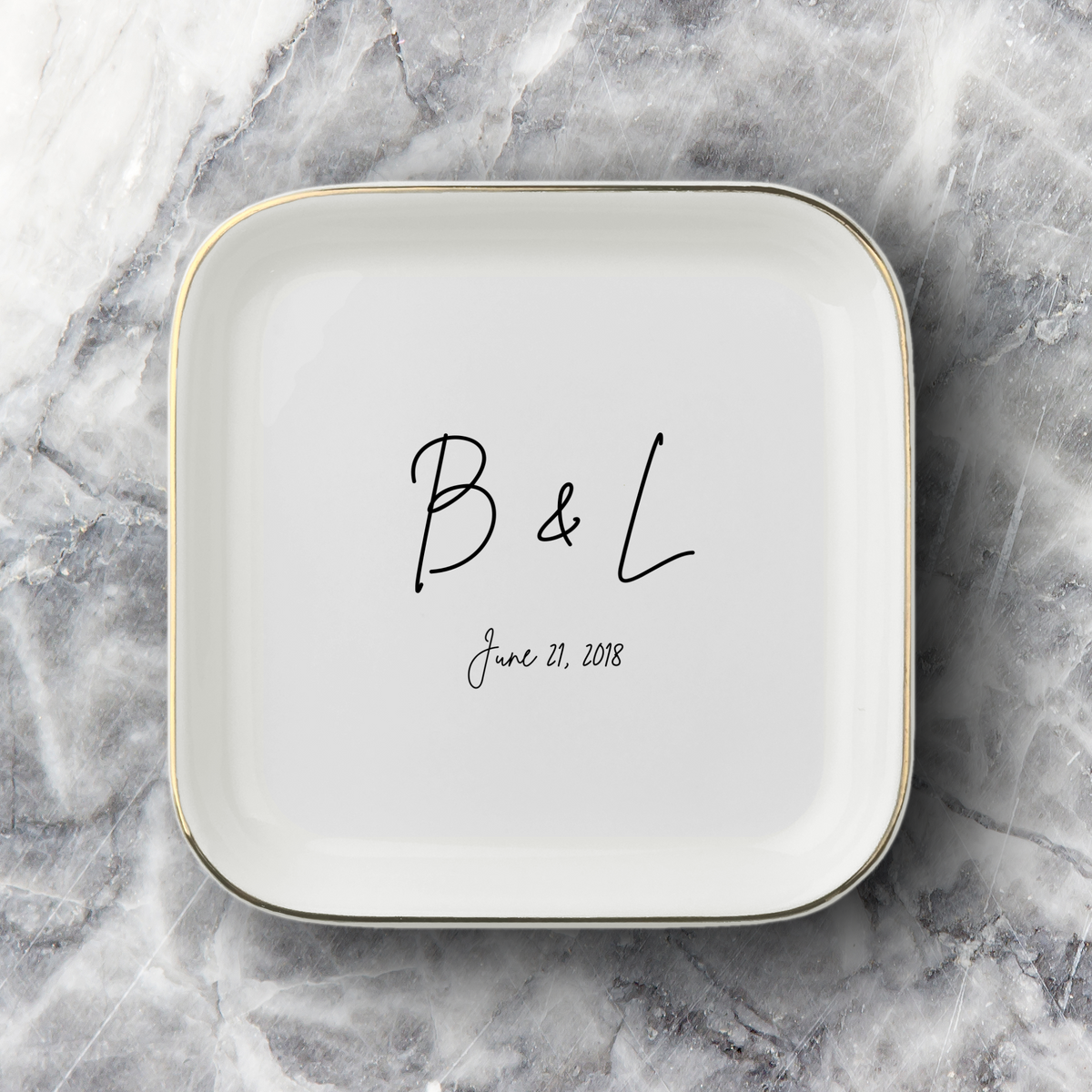 A square white ceramic dish with rounded corners, featuring a thin gold rim and personalized black script text reading 'B & L' and the date 'June 21, 2018', set on a gray and white marble surface.